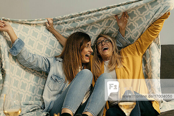 Cheerful daughter and mother holding blanket on sofa in living room