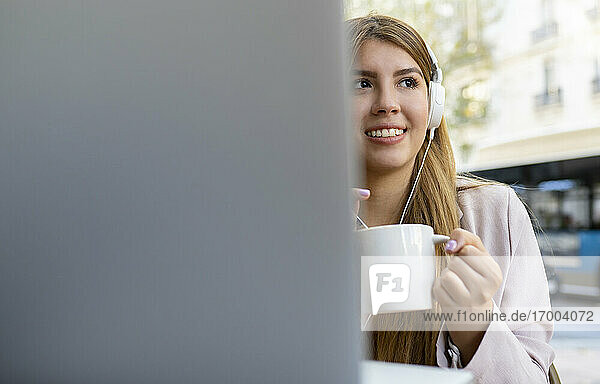 Lächelnde junge Frau mit Laptop und Kaffeetasse  die wegschaut  während sie in einem Straßencafé sitzt