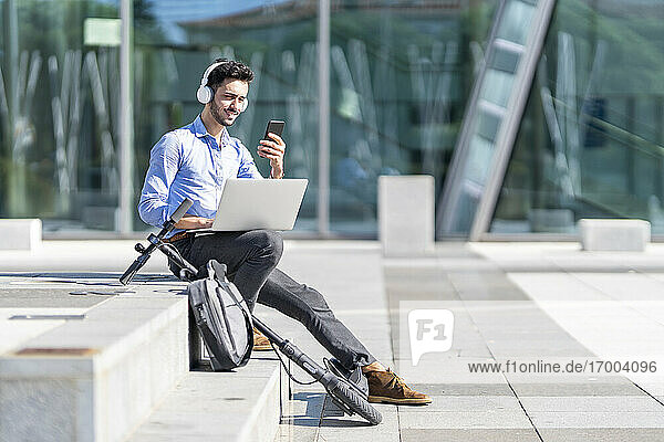 Geschäftsmann mit Kopfhörern  der ein Mobiltelefon benutzt  während er mit einem Laptop auf einem Elektroroller und einer Aktentasche auf einer Treppe sitzt