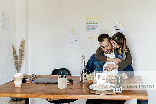 Lächelnde weibliche Fachkraft  die einen männlichen Mitarbeiter umarmt  während sie im Büro zu Hause isst