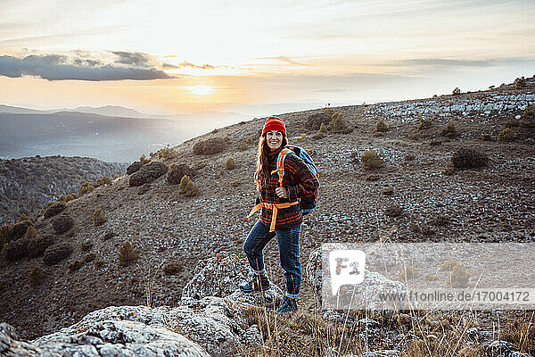 Lächelnde Wanderin  die wegschaut  während sie bei Sonnenuntergang auf einem Felsen steht