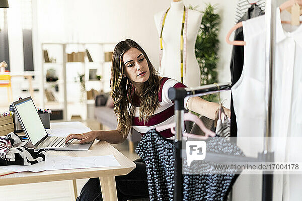 Junge Geschäftsfrau mit Laptop betrachtet Tops  die auf einem Gestell in einem Studio angeordnet sind