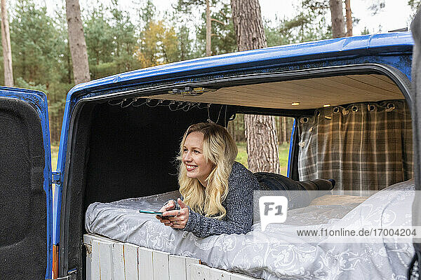 Glückliche Frau mit Handy träumt  während sie auf dem Bett im Van liegt