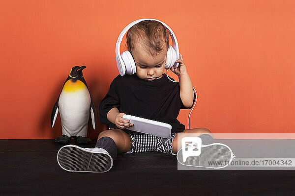 Baby-Mädchen mit Kopfhörern  das ein Mobiltelefon benutzt  während es neben einem Spielzeugpinguin vor einem orangefarbenen Hintergrund sitzt
