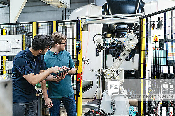 Male coworkers holding digital tablet looking at robotic arm while standing in manufacturing industry