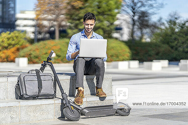 Geschäftsmann  der an einem Laptop arbeitet  während er mit einer Aktentasche und einem elektrischen Motorroller in der Stadt sitzt