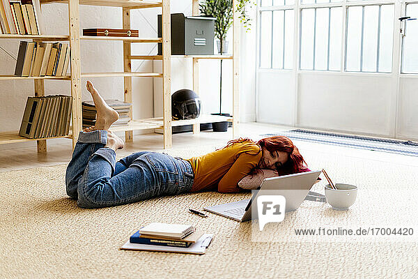 Rothaarige Frau mit Buch und Laptop auf Teppich liegend zu Hause