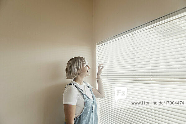 Glückliche junge Frau  die durch Jalousien auf das Fenster schaut