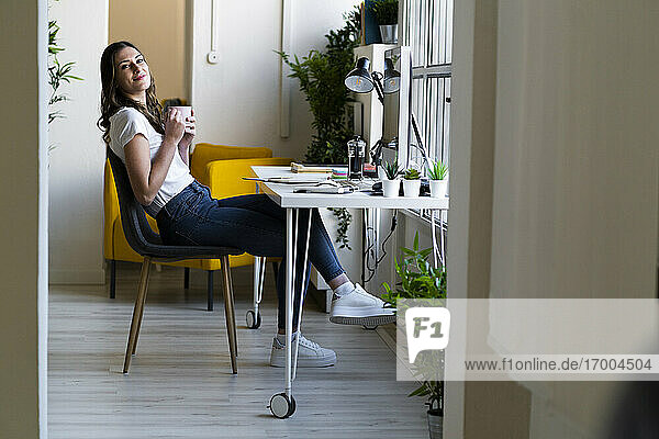 Lächelnde Geschäftsfrau  die eine Kaffeetasse hält und auf einem Stuhl im Büro sitzt