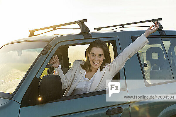 Unbekümmerte junge Frau lehnt sich bei Sonnenuntergang mit erhobenen Armen aus dem Autofenster