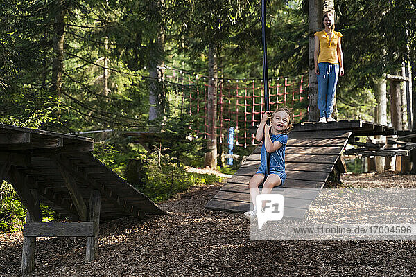 Kleines Mädchen reitet auf einer Seilrutsche im Wald  die Mutter steht im Hintergrund
