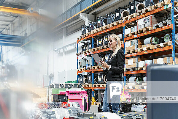 Blond female entrepreneur holding digital tablet looking away while standing in factory