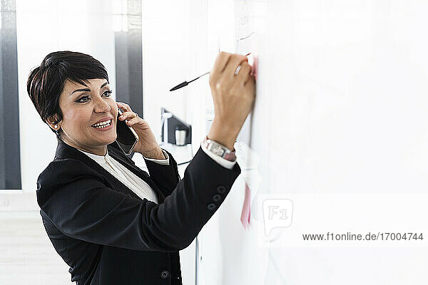 Portrait of businesswoman talking on smart phone and writing on adhesive note hanging on office whiteboard