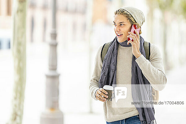 Lächelnder Mann mit Kaffeetasse  der beim Spazierengehen in der Stadt mit seinem Mobiltelefon spricht