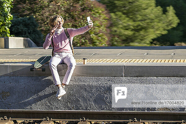 Lächelnder Mann gestikuliert Friedenszeichen  während er ein Selfie mit Longboard auf dem Bahnsteig macht
