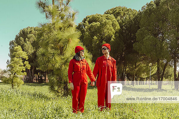 Junges Paar in roten Overalls und Hüten steht Hand in Hand auf einer Wiese und sieht sich an