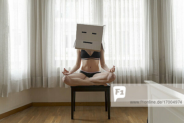 Frau mit Karton auf dem Kopf und gelangweiltem Smiley sitzt auf einem Stuhl