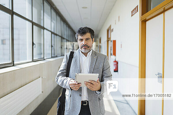 Reifer männlicher Professor  der ein digitales Tablet in einem Korridor einer Universität hält