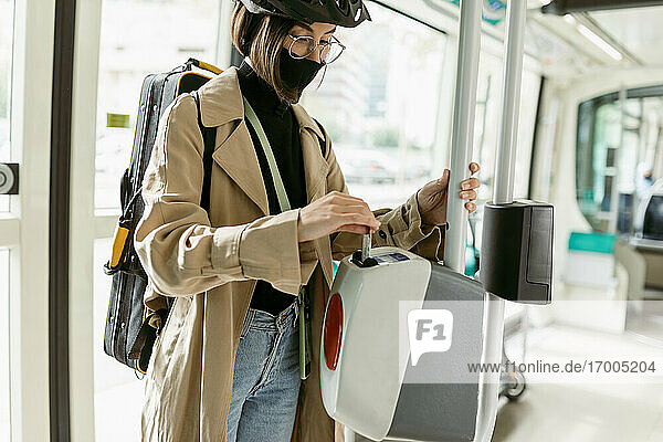 Frau mit Gesichtsmaske und Fahrradhelm  die in der Straßenbahn eine Fahrkarte in den Entwertungsautomaten steckt
