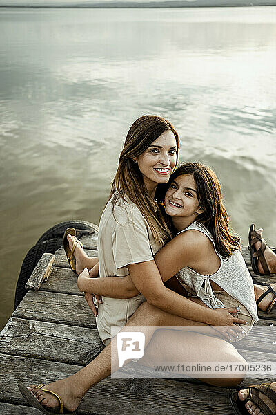 Mother and daughter embracing each other while sitting at jetty against lake