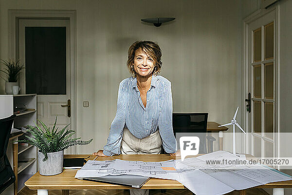 Lächelnde Architektin  die sich im Stehen auf den Schreibtisch stützt  im Büro