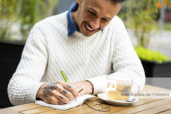 Lächelnder Mann schreibt in ein Buch  während er in einem Straßencafé eine Teepause macht