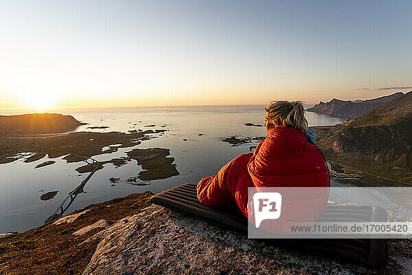 Frau in Schlafsack eingewickelt mit Blick auf die Aussicht
auf dem Berg Volandstinden sitzend  Lofoten  Norwegen