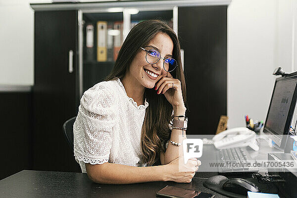 Lächelnde Geschäftsfrau sitzt mit dem Kopf in der Hand im Büro