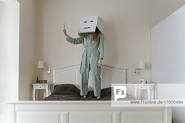 Frau  die eine Pappschachtel auf dem Kopf mit einem gelangweilten Smiley trägt und ein Selfie auf ihrem Bett macht