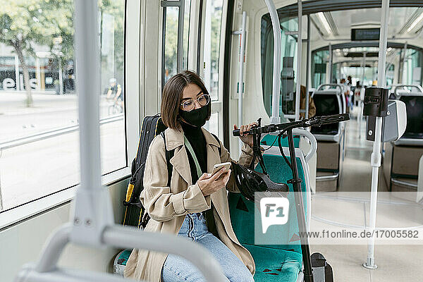 Mittlere erwachsene Frau mit Gesichtsmaske  die ein Mobiltelefon benutzt  während sie mit einem Elektroroller in der Straßenbahn sitzt