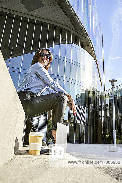 Lächelnde Geschäftsfrau sitzt auf einer Bank neben einem Bürogebäude