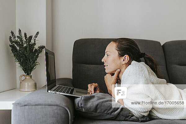 Frau benutzt einen Laptop  während sie auf dem Sofa im Wohnzimmer liegt