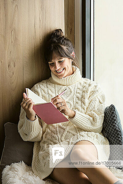Smiling woman writing in book while sitting by window at home