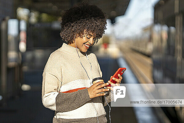 Lächelnde junge Frau  die ein Mobiltelefon benutzt  während sie am Bahnhof Musik hört