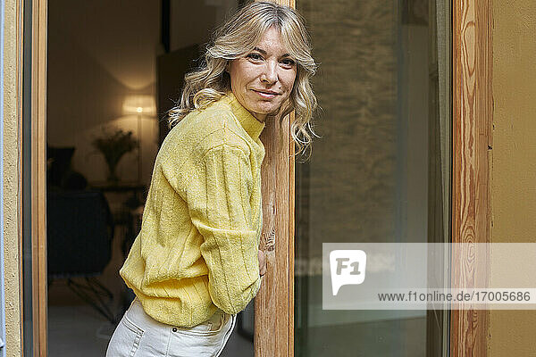 Lächelnde reife Frau mit blondem Haar lehnt sich an die Tür zu Hause