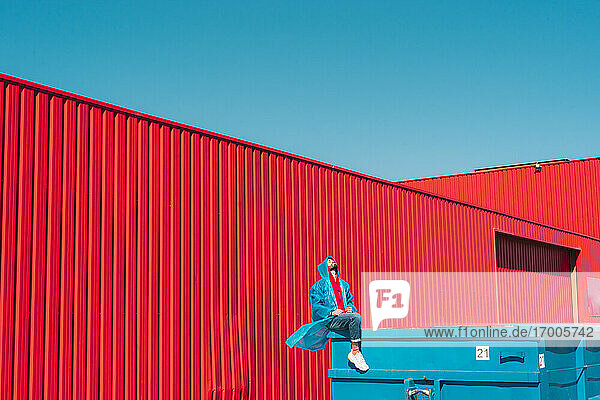 Junger Mann in Regenjacke sitzt auf dem Rand eines blauen Containers vor einer roten Wand und schaut nach oben