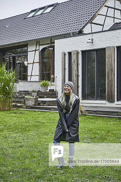 Lachende reife Frau im Regenmantel  die im Gras vor einem Haus steht