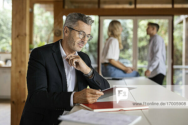Geschäftsmann  der ein digitales Tablet benutzt  während er zu Hause an einem Tisch mit Kollegen im Hintergrund steht