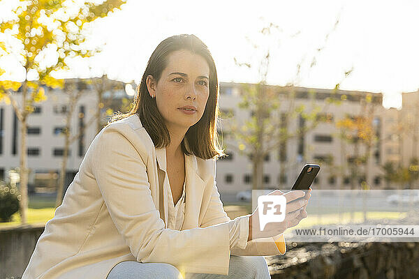 Businesswoman holding smart phone looking away while sitting in city
