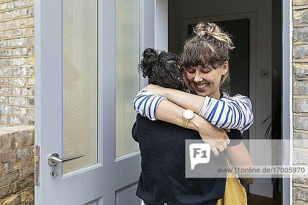 Smiling woman embracing girlfriend while standing at home entrance door