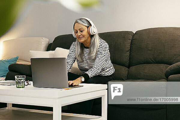Lächelnde Frau mit Kopfhörern und Laptop  während sie zu Hause auf dem Sofa sitzt