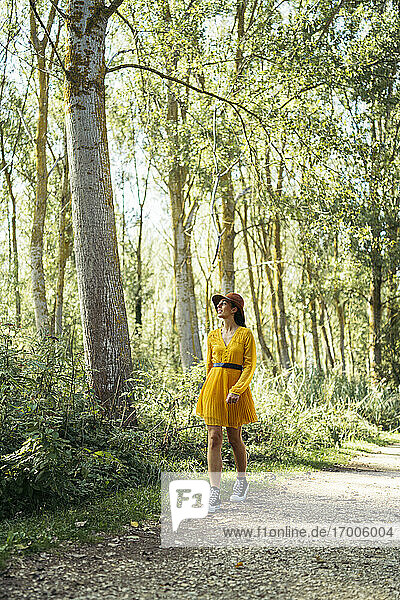 Young woman wearing brown hat  yellow dress and black sneakers looking up and walking in forest