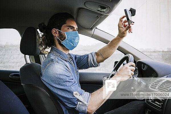 Young man wearing protective face mask while adjusting mirror in car