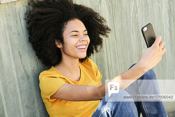 Smiling student taking selfie through mobile phone while sitting against wall