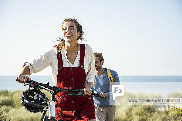 Lächelnde Freundin hält Fahrräder beim Spaziergang mit Freund gegen das Meer