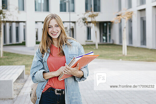 Lächelnde schöne blonde junge Frau hält Bücher auf dem Universitätscampus