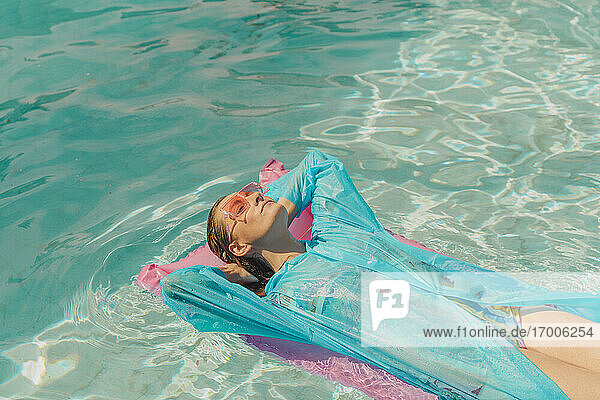 Woman wearing blue rain coat relaxing on pink airbed in swimming pool