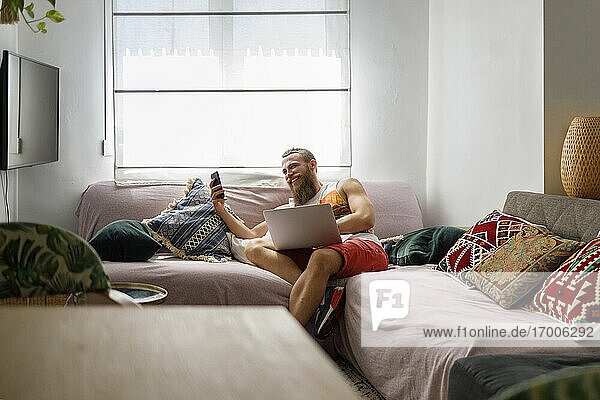 Lächelnder Mann  der sein Smartphone benutzt  während er mit seinem Laptop auf dem Sofa im Wohnzimmer sitzt