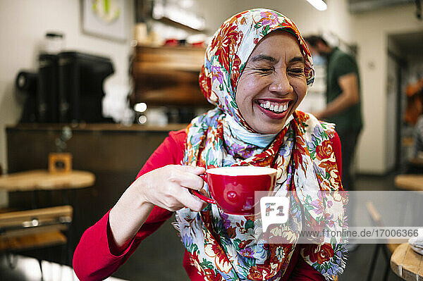Frau mit geblümtem Hidschab lacht  während sie eine Kaffeetasse in einem Café hält