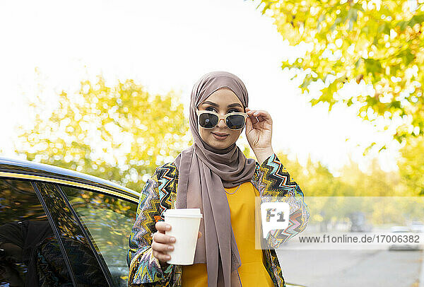 Frau mit Kaffeetasse und Sonnenbrille neben einem Auto stehend